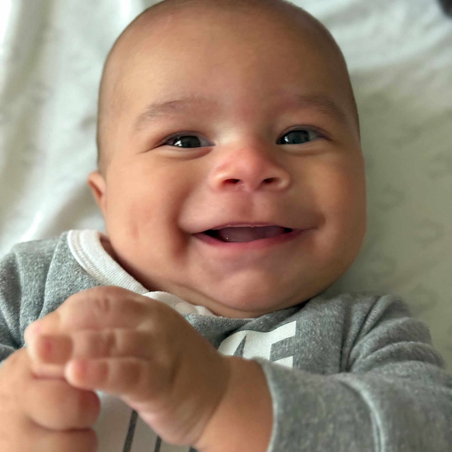 Matheus is registered to the contest to win money with this photo: baby, smiling, close_up, infant, hands, clapping, gray_clothing, cheeks, happy, face, portrait, young_child, cute, indoor, blanket, soft_light, expression, head, skin, person