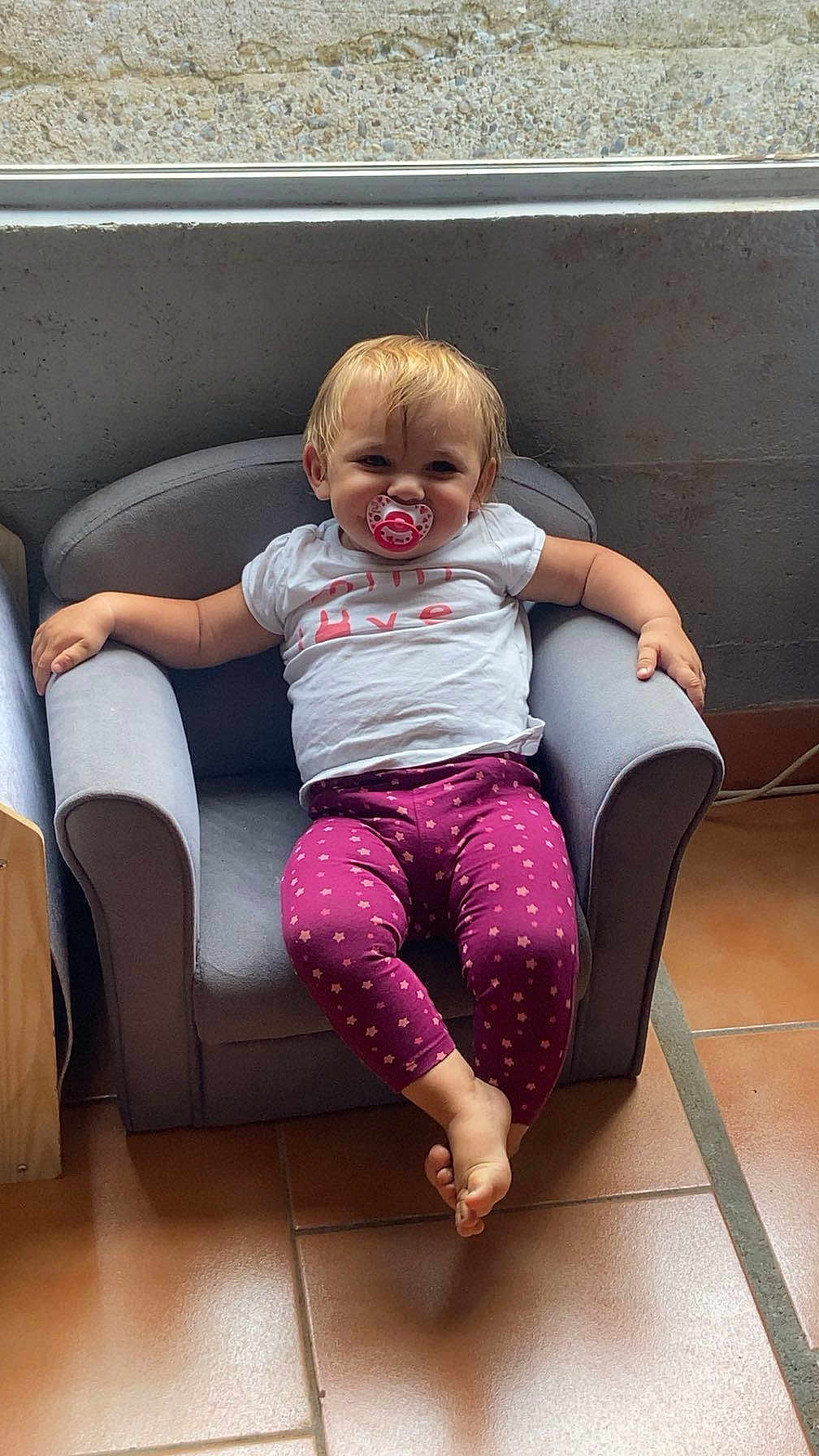 Léna a rejoint le concours — aidez-le/la à gagner de superbes lots ! toddler, child, pacifier, purple_leggings, white_tshirt, armchair, indoor, tile_floor, relaxed, smiling, blond_hair, furniture, person, casual, seated, small_chair, cute, baby, home, portrait
