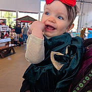 Riley joined the competition — help win amazing prizes! baby, toddler, child, smiling, blue_eyes, red_bow, headband, velvet_dress, green_dress, hand_in_mouth, indoor, market, crowd, holiday_decor, string_lights, portrait, cheerful, being_held, window_light, blurred_background