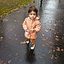 autumn_leaves, casual_clothing, child, curious, daytime, fall, footwear, outdoor, park, pavement, person, raincoat, seasonal, small_toy, street, toddler, toy, walking_path, wet_pavement, young_child