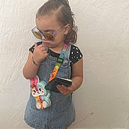Nashley is registered to the contest to win money with this photo: child, toddler, girl, portrait, standing, sunglasses, tablet, overalls, dress, star_pattern, hair_bow, ponytail, stuffed_toy, unicorn, necklace, bracelet, sandals, indoor, textured_wall, tile_floor