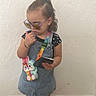 child, toddler, girl, portrait, standing, sunglasses, tablet, overalls, dress, star_pattern, hair_bow, ponytail, stuffed_toy, unicorn, necklace, bracelet, sandals, indoor, textured_wall, tile_floor