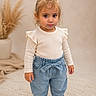 child, toddler, girl, portrait, standing, smiling, fashion, clothing, sweater, ruffles, jeans, sneakers, bow, indoors, studio, rug, plant, home_decor, blonde_hair, photography