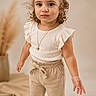 child, girl, toddler, portrait, standing, curly_hair, smiling, fashion, white_top, beige_trousers, sneakers, necklace, indoor, studio, neutral_background, home_decor, pampas_grass, full_body, photography, fashionable
