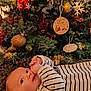 baby, infant, christmas_tree, ornaments, holiday, decorations, tinsel, blanket, striped_clothing, festive, indoors, cute, soft_texture, gaze, hands, face, celebration, warm_lighting, seasonal, holiday_spirit