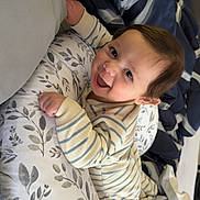 Roman is registered to the contest to win money with this photo: baby, bed, blanket, child, cozy, cute, face, floral_pattern, happy, indoors, infant, lying_down, person, pillow, playful, resting, sleepwear, smiling, striped_pajamas, young_child