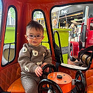 Lisandro a rejoint le concours — aidez-le/la à gagner de superbes lots ! bear_pattern, child, cute, glasses, indoor, orange, person, play, portrait, red, seat, seatbelt, steering_wheel, storefront, sweater, toddler, toy, urban, vehicle, window