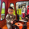 bear_pattern, child, cute, glasses, indoor, orange, person, play, portrait, red, seat, seatbelt, steering_wheel, storefront, sweater, toddler, toy, urban, vehicle, window
