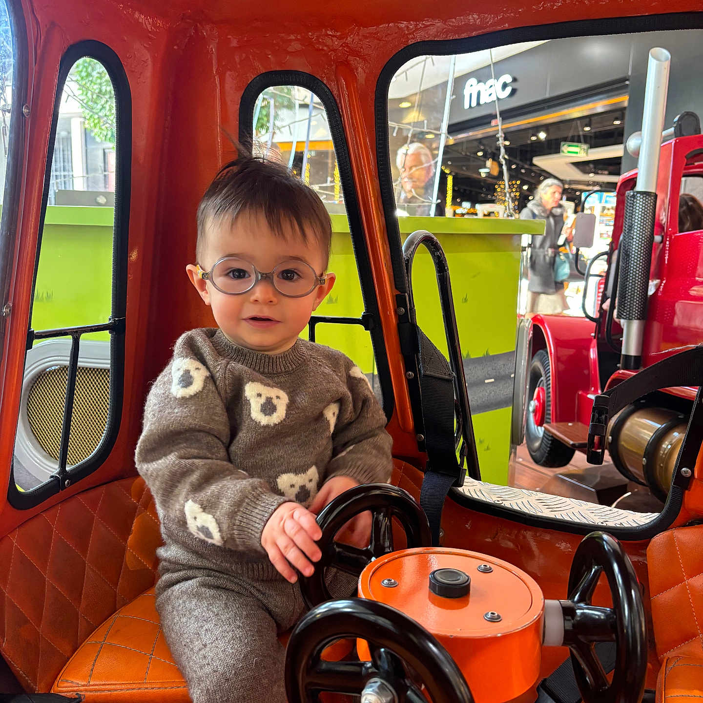Lisandro a rejoint le concours — aidez-le/la à gagner de superbes lots ! bear_pattern, child, cute, glasses, indoor, orange, person, play, portrait, red, seat, seatbelt, steering_wheel, storefront, sweater, toddler, toy, urban, vehicle, window