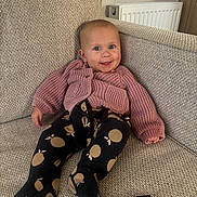 Maeve is registered to the contest to win money with this photo: baby, child, couch, cozy, cute, face, footed_pants, furniture, happy, home, indoor, infant, knitwear, pants, person, relaxed, remote_control, seated, smile, sweater