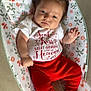 baby, infant, curly_hair, red_pants, white_shirt, floral_pattern, cushion, bare_feet, infant_clothing, lying_down, indoor, cute, child, portrait, small_hands, small_feet, expression, comfort, soft_fabric, casual