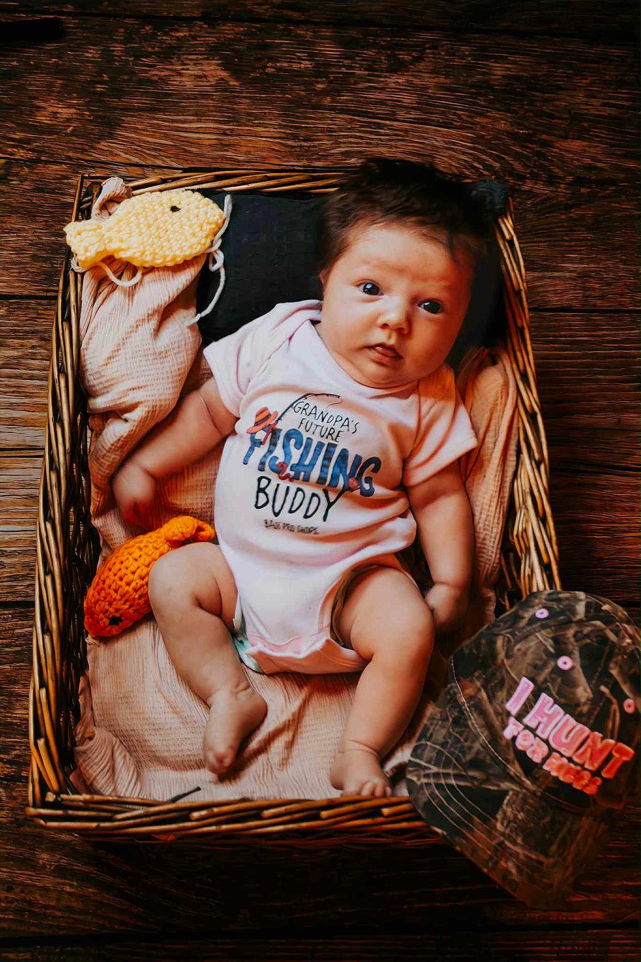 Gracelynn is registered to the contest to win money with this photo: baby, basket, blanket, wood_floor, onesie, knitted_fish, camo_hat, infant, lying_down, cozy, indoor, cute, child, person, soft_texture, plush, newborn, portrait, clothing, resting