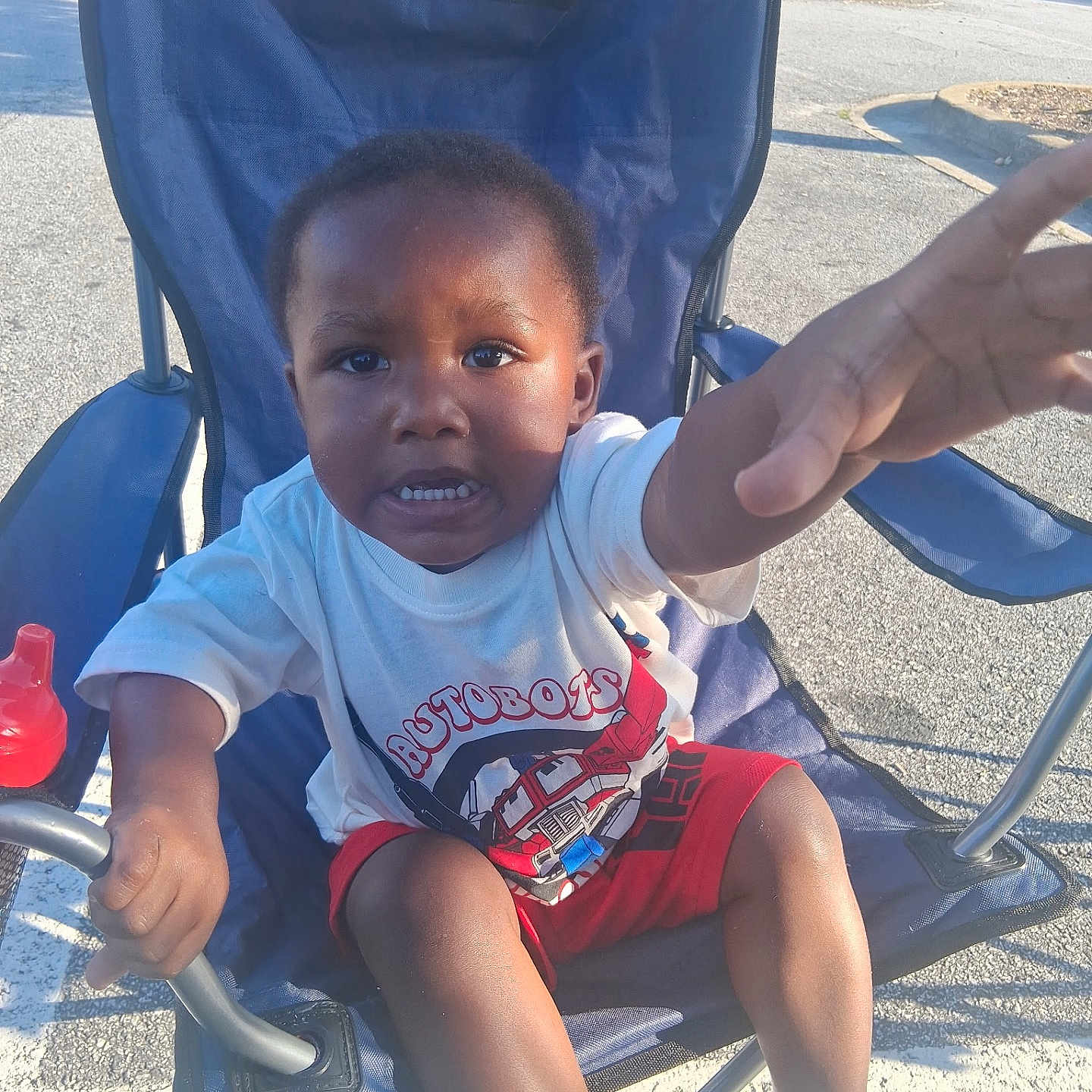 Jeremiah joined the competition — help win amazing prizes! child, toddler, boy, chair, outdoor, summer, clothing, shirt, shorts, footwear, flip_flops, hand, face, expression, sunlight, pavement, parking_lot, toy, sippy_cup, determined