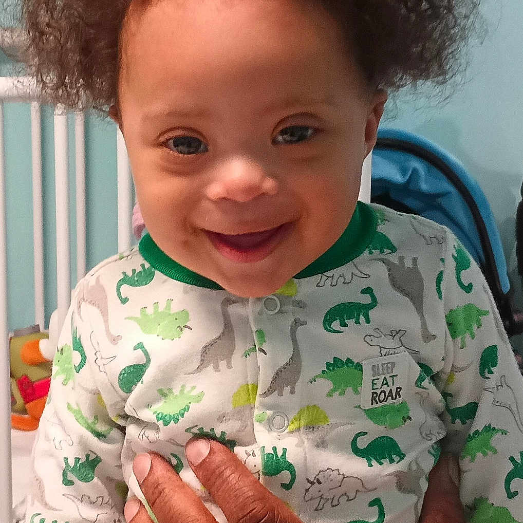 Elijah is registered to the contest to win money with this photo: adult_hand, baby, blue_wall, child, closeup, clothing, crib, curly_hair, cute, dinosaur_print, hand, happy, indoor, infant, onesie, person, portrait, smiling, supporting_hand, toddler