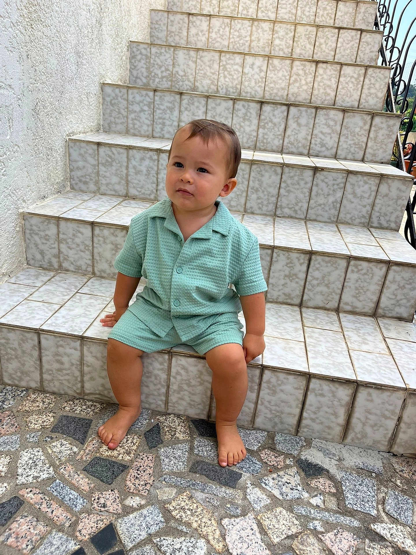 Manua a rejoint le concours — aidez-le/la à gagner de superbes lots ! toddler, child, steps, tile, barefoot, outdoor, curious, green_clothing, mosaic_floor, sitting, person, young_child, texture, pattern, casual, daylight, concrete, wall, portrait, expression