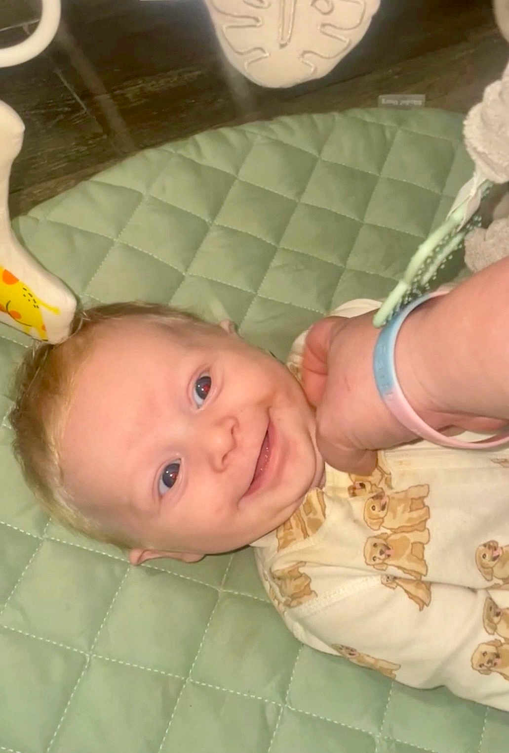 Virgil is registered to the contest to win money with this photo: baby, infant, smiling, onesie, dog_prints, hand, wristband, quilted_mat, green_mat, indoor, cute, happy, child, face, person, lying_down, closeup, skin, head, expression