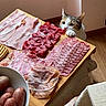 cat, curious, deli_meat, sliced_meat, wooden_board, table, indoor, food, snack, paw, pink_meat, salami, prosciutto, ham, kitchen, pet, animal, floor, curiosity, appetizer