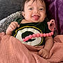 baby, child, tongue_out, blanket, pacifier, clothing, smile, happy, face, person, indoors, cute, wrapped, pink, green, logo, hand, portrait, infant, cozy