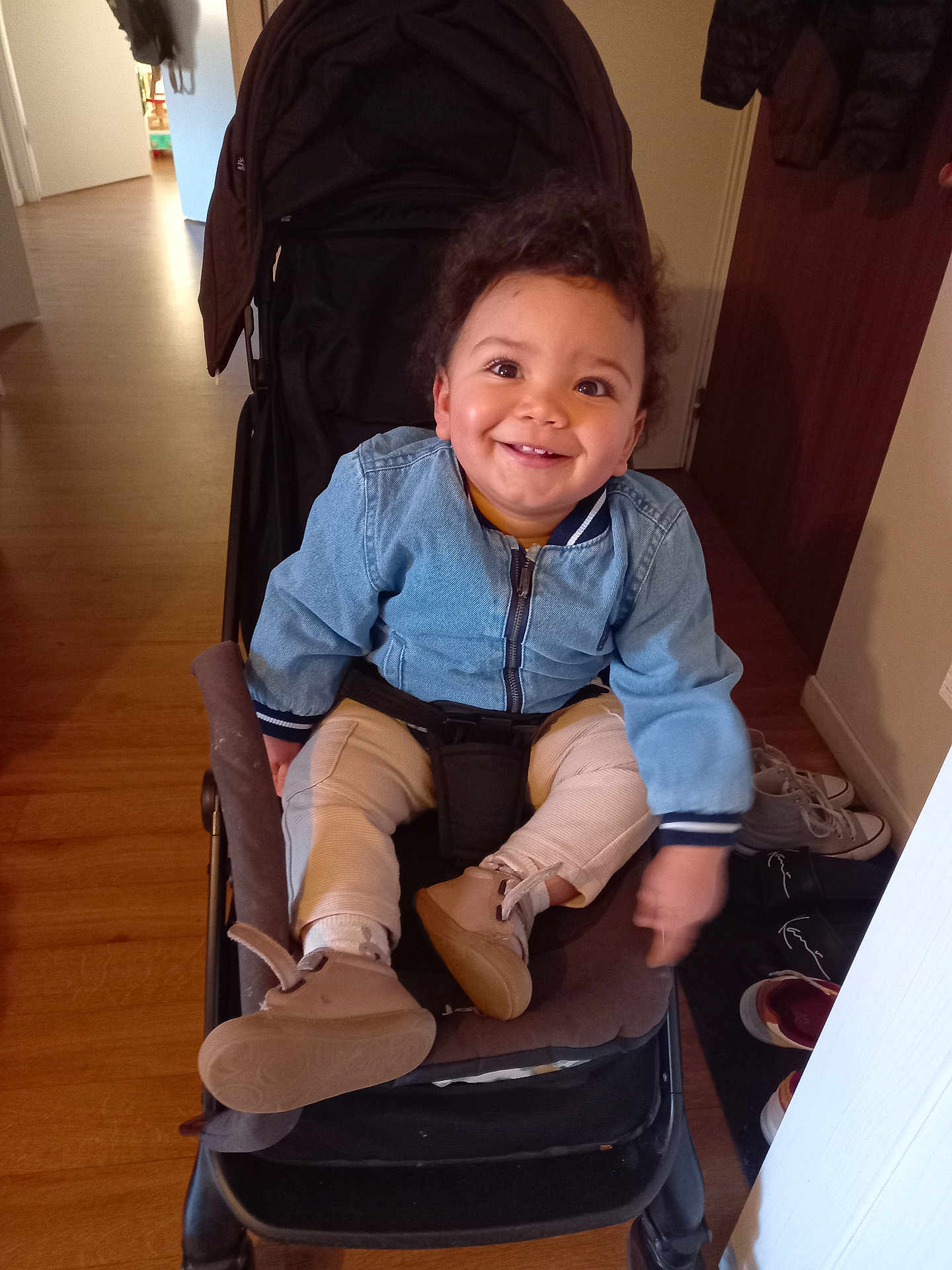 Gabriel a rejoint le concours — aidez-le/la à gagner de superbes lots ! child, toddler, baby, stroller, smiling, indoor, jean_jacket, curly_hair, brown_shoes, socks, wooden_floor, hallway, doorway, coat, seat_belt, portrait, happy, sneakers, shoe_rack, stroller_seat