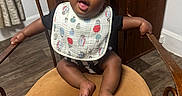 Atlas is registered to the contest to win money with this photo: baby, child, infant, sitting, high_chair, wooden_chair, bib, smiling, tongue_out, indoor, portrait, dark_skin, black_hair, braids, arms_outstretched, bare_feet, wood_floor, furniture, home, happy