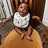 baby, child, infant, sitting, high_chair, wooden_chair, bib, smiling, tongue_out, indoor, portrait, dark_skin, black_hair, braids, arms_outstretched, bare_feet, wood_floor, furniture, home, happy