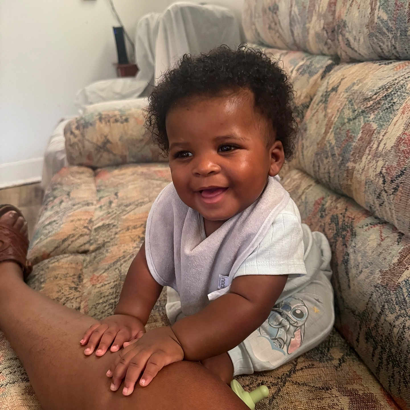 Atlas is registered to the contest to win money with this photo: baby, child, infant, smiling, happy, portrait, curly_hair, sitting, couch, sofa, living_room, indoor, home, adult_leg, clothing, bib, pajamas, pacifier, furniture, photography