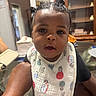 baby, child, infant, portrait, face, eyes, curious, indoors, home, bib, clothing, hair_buns, sitting, being_held, adult_arm, furniture, cabinet, dining_room, smiling, photography