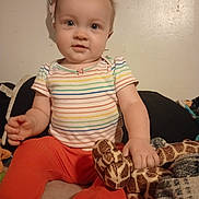 Willow joined the competition — help win amazing prizes! baby, child, plush_toy, giraffe, striped_shirt, orange_pants, indoor, bed, blanket, comfort, cute, smile, person, young_child, sitting, toy, soft_toy, home, cozy, portrait