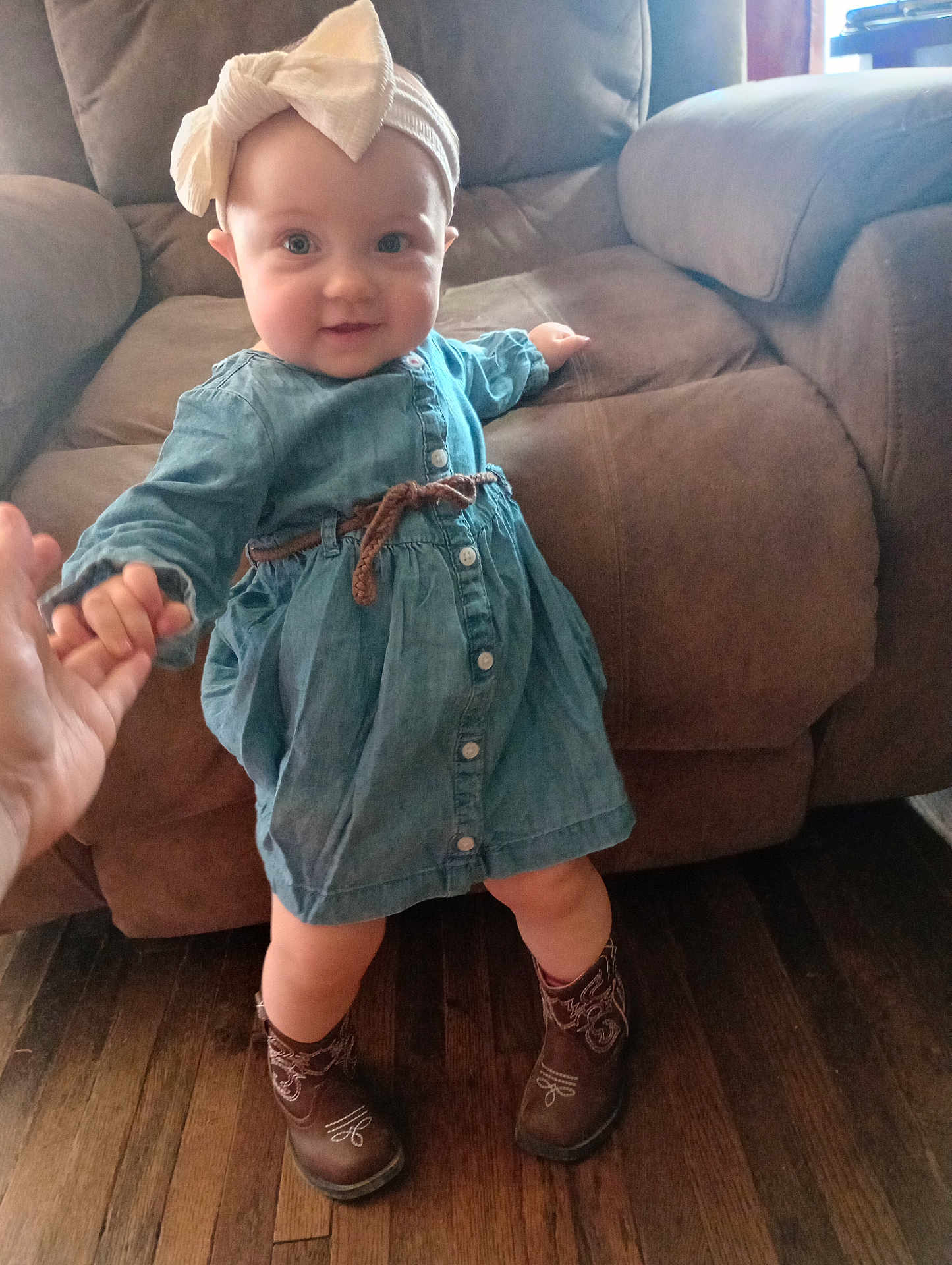 Willow is registered to the contest to win money with this photo: baby, child, denim_dress, bow_headband, cowboy_boots, hand_holding, wooden_floor, couch, indoor, smiling, person, face, footwear, clothing, furniture, happy, standing, cute, support, home