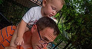 Chaos is registered to the contest to win money with this photo: bonding, casual_clothing, child_on_shoulders, curious, daylight, family, fence, glasses, greenery, head_and_shoulders, looking_down, man, nature, orange_shirt, outdoor, person, portrait, toddler, trees, young_child