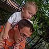 bonding, casual_clothing, child_on_shoulders, curious, daylight, family, fence, glasses, greenery, head_and_shoulders, looking_down, man, nature, orange_shirt, outdoor, person, portrait, toddler, trees, young_child