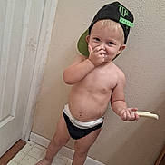 Chaos joined the competition — help win amazing prizes! backward_hat, barefoot, cap, child, diaper, door, expression, face, floor, food, hand, indoor, light_skin, mischievous, playful, standing, tile_floor, toddler, wall, young_child