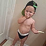 backward_hat, barefoot, cap, child, diaper, door, expression, face, floor, food, hand, indoor, light_skin, mischievous, playful, standing, tile_floor, toddler, wall, young_child