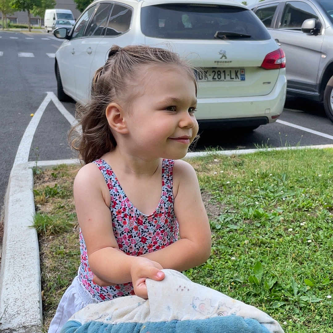 Leïla participe au concours pour gagner de l'argent avec cette photo : blanket, car, casual_clothing, child, curly_hair, daytime, grass, greenery, happy, holding, nature, outdoor, parking_lot, person, playful, shorts, side_view, sleeveless_top, smiling, young