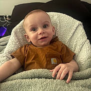 Tony joined the competition — help win amazing prizes! baby, infant, blanket, brown_shirt, cozy, smiling, face, hand, bedroom, pillow, child, cute, indoors, portrait, soft_texture, resting, young_child, comfortable, expression, toddler