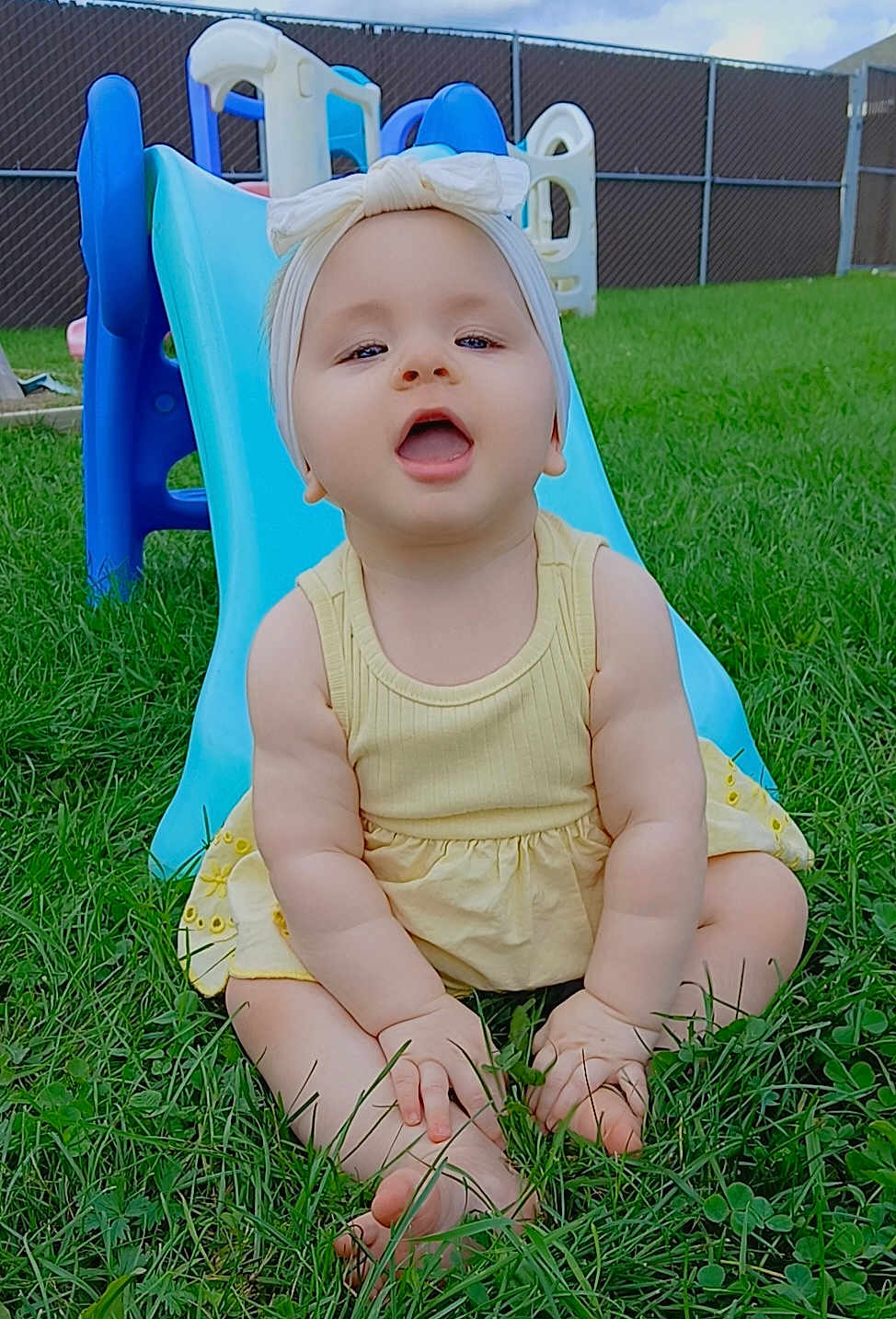 Blessing joined the competition — help win amazing prizes! baby, child, grass, slide, playground, outdoor, yellow_dress, headband, smiling, happy, barefoot, summer, toy, fence, blue_slide, infant, cute, sitting, nature, daytime