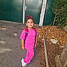 child, girl, pink_clothing, backpack, outdoor, path, autumn_leaves, green_gate, tree, sidewalk, casual, smiling, nature, daylight, footwear, hair_tie, standing, curly_hair, playful, park