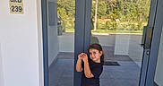 Mady participe au concours pour gagner de l'argent avec cette photo : child, glass_door, reflection, outdoor, building_entrance, tile_floor, black_tshirt, beige_pants, sneakers, mat, trees, daylight, posing, smiling, person, modern_architecture, window, door_handle, wall, sign