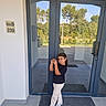 child, glass_door, reflection, outdoor, building_entrance, tile_floor, black_tshirt, beige_pants, sneakers, mat, trees, daylight, posing, smiling, person, modern_architecture, window, door_handle, wall, sign