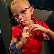 Bryleigh joined the competition — help win amazing prizes! child, girl, glasses, red_dress, scrunchie, hand_gesture, heart_shape, seat, indoor, portrait, blonde_hair, casual_clothing, expression, puckered_lips, young, person, eyewear, blue_scrunchie, dim_lighting, window
