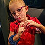 child, girl, glasses, red_dress, scrunchie, hand_gesture, heart_shape, seat, indoor, portrait, blonde_hair, casual_clothing, expression, puckered_lips, young, person, eyewear, blue_scrunchie, dim_lighting, window
