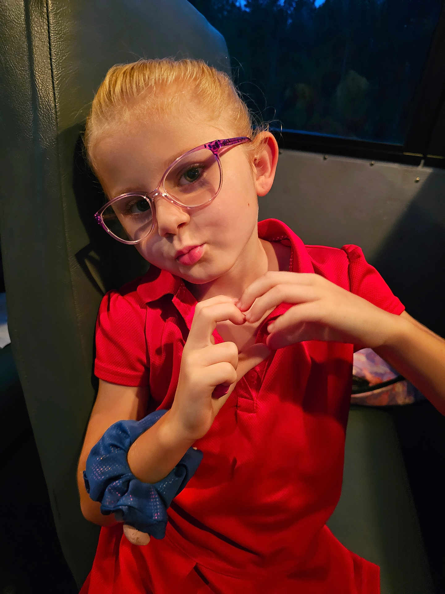 Bryleigh joined the competition — help win amazing prizes! child, girl, glasses, red_dress, scrunchie, hand_gesture, heart_shape, seat, indoor, portrait, blonde_hair, casual_clothing, expression, puckered_lips, young, person, eyewear, blue_scrunchie, dim_lighting, window