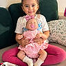 child, girl, toddler, doll, baby_doll, smiling, portrait, sitting, couch, sofa, pink_cushion, pink_clothing, white_shirt, bow, pacifier, blanket, pillow, indoor, playtime, family_home