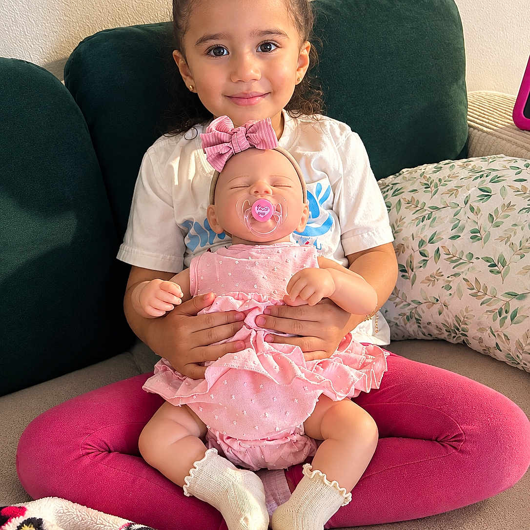 Karla joined the competition — help win amazing prizes! child, girl, toddler, doll, baby_doll, smiling, portrait, sitting, couch, sofa, pink_cushion, pink_clothing, white_shirt, bow, pacifier, blanket, pillow, indoor, playtime, family_home