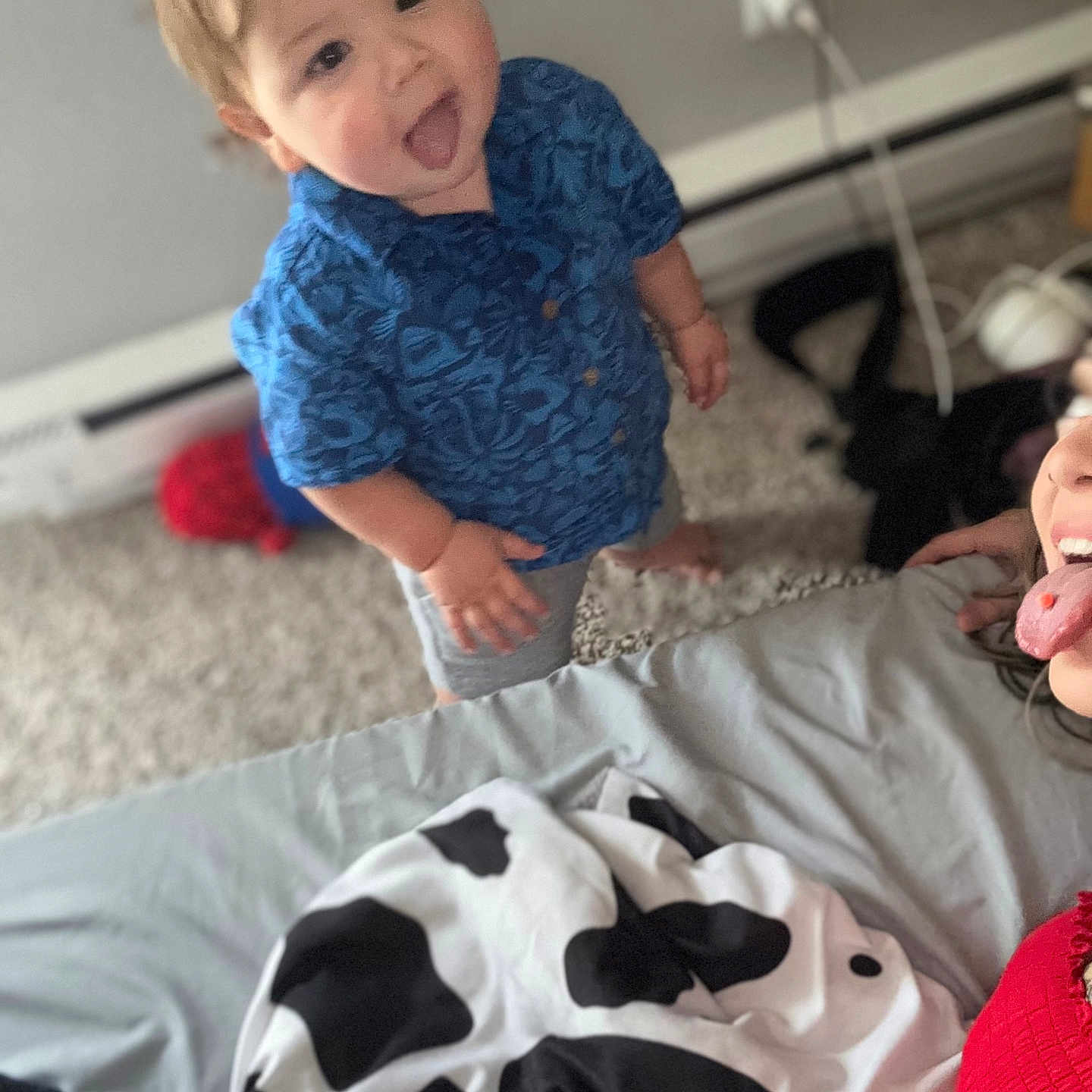 Jaymin is registered to the contest to win money with this photo: adult, bed, blanket, blue_shirt, candid, carpet, charger, child, fun, home, indoor, patterned_shirt, person, person_standing, playful, power_outlet, red_clothing, smiling, toddler, tongue_out