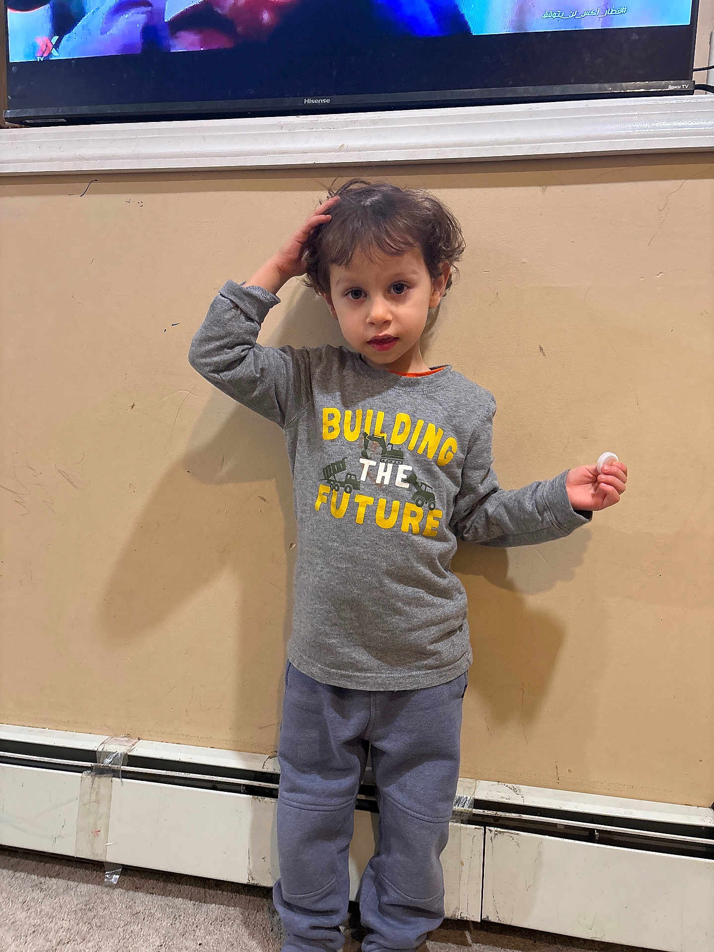 Yamen M joined the competition — help win amazing prizes! toddler, child, indoor, standing, grey_shirt, yellow_text, curly_hair, wall, carpet, pants, hand, face, expression, holding_object, casual_clothing, home_interior, radiator, curious, young_child, portrait
