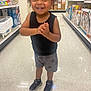 toddler, child, smiling, standing, clapping_hands, store, aisle, toys, baby_products, sneakers, shorts, black_shirt, curly_hair, happy, floor, shelf, retail, shopping, indoor, person