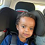 child, toddler, car_seat, curly_hair, blue_shirt, sippy_cup, seatbelt, car_interior, window, natural_light, face, eyes, smile, young_child, safety_harness, headrest, person, portrait, indoors, casual_clothing