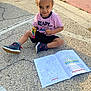 child, toddler, sitting, pavement, coloring_book, toy, striped_shirt, shorts, sneakers, outdoor, sidewalk, sunlight, happy, smiling, playful, person, clothing, casual, young, recreation