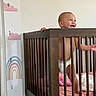 baby, crib, wooden_crib, smiling, standing, height_chart, rainbow, clouds, nursery, diaper, pink_bedding, toy, baby_room, child, infant, happy, indoor, furniture, person, home